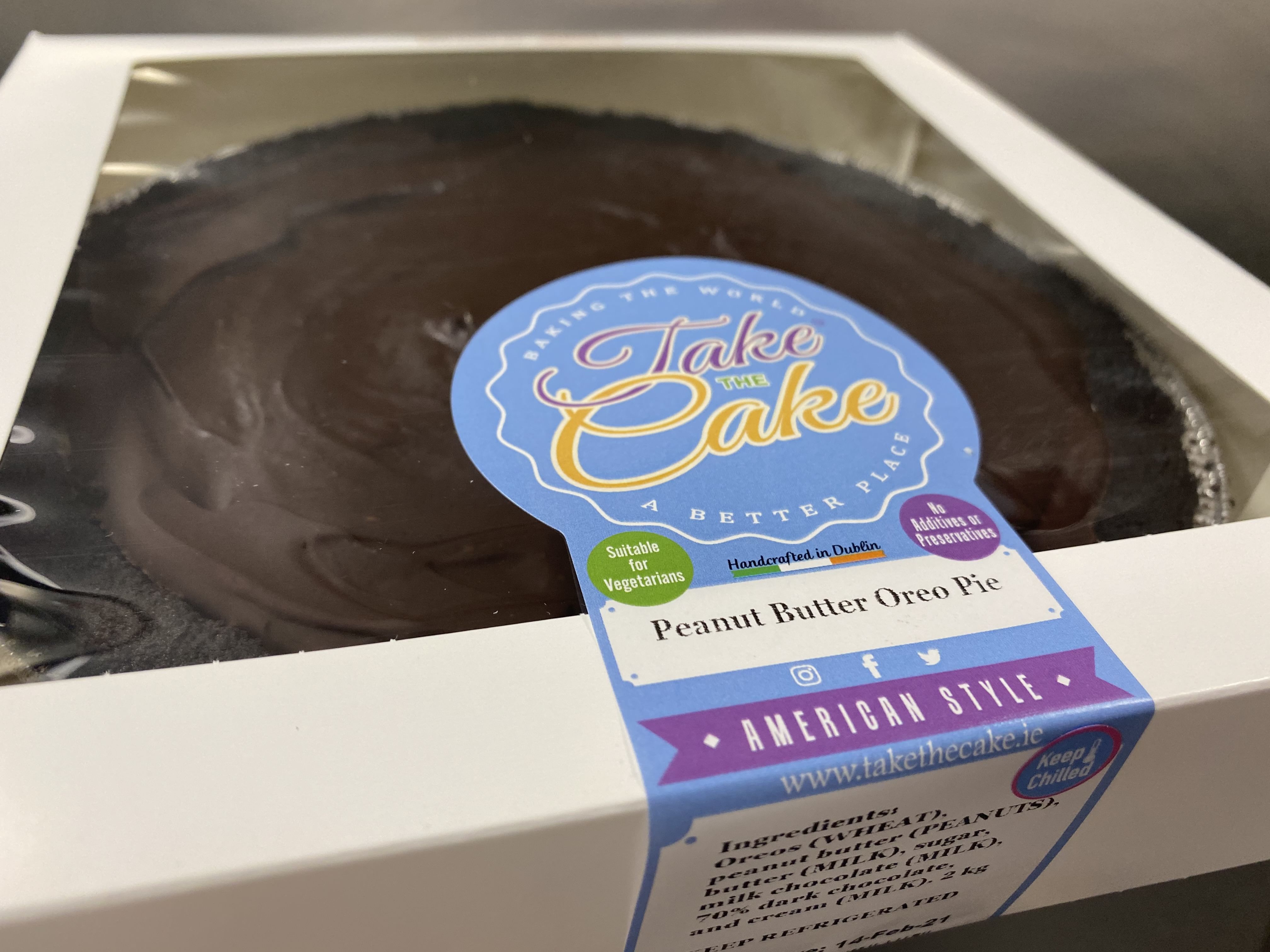 A close-up image of a boxed dessert labeled 'Peanut Butter Oreo Pie' with a large blue logo on the packaging reading 'Take the Cake.' The packaging indicates the product is suitable for vegetarians and is handcrafted in Dublin. The pie has a rich chocolate ganache topping and creamy peanut butter filling.